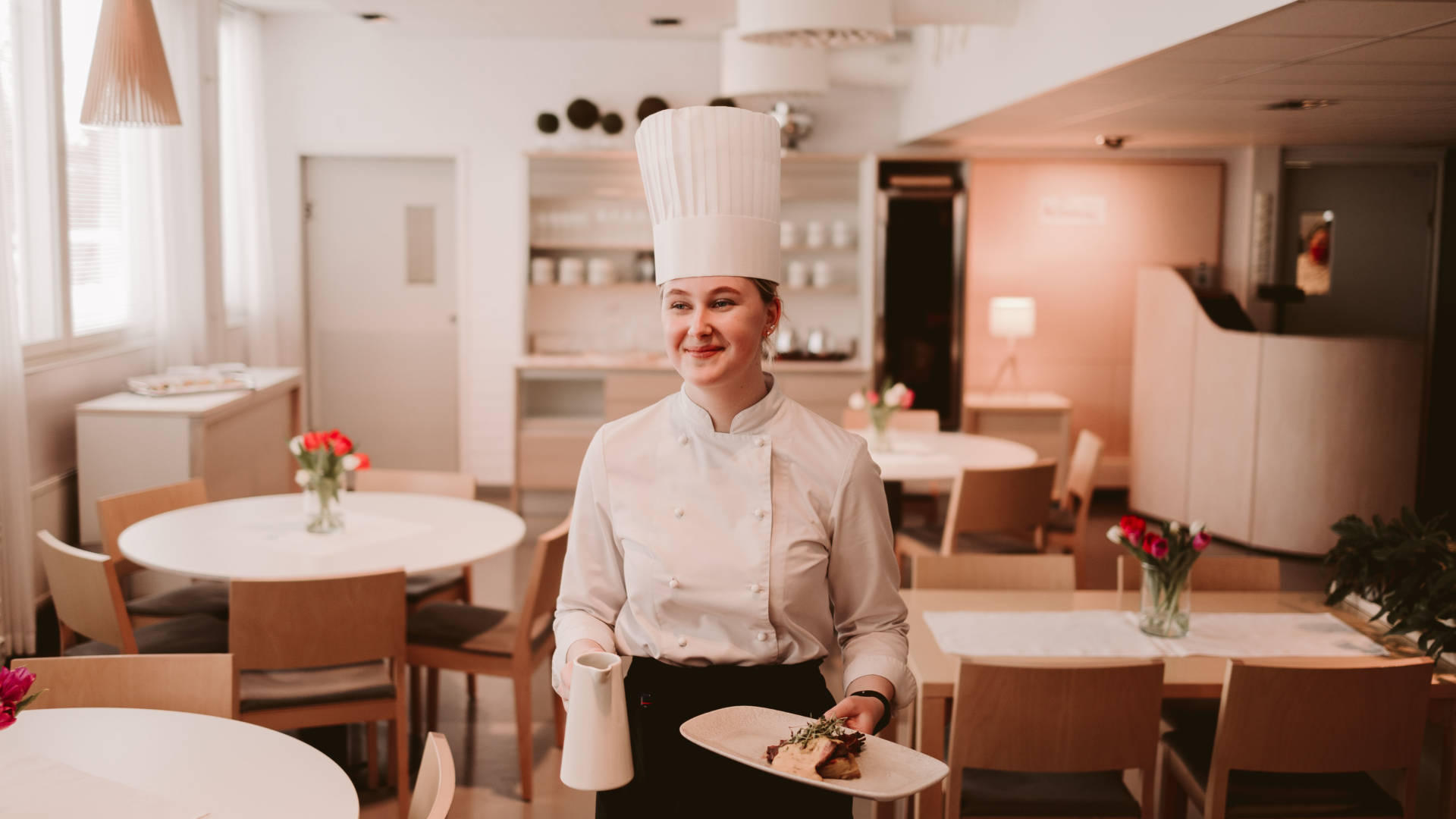 Nuori iloinen ravintola-alan opiskelija tarjoilee ruoka annosta opetusravintola Poukamassa kokin päähine päässään.